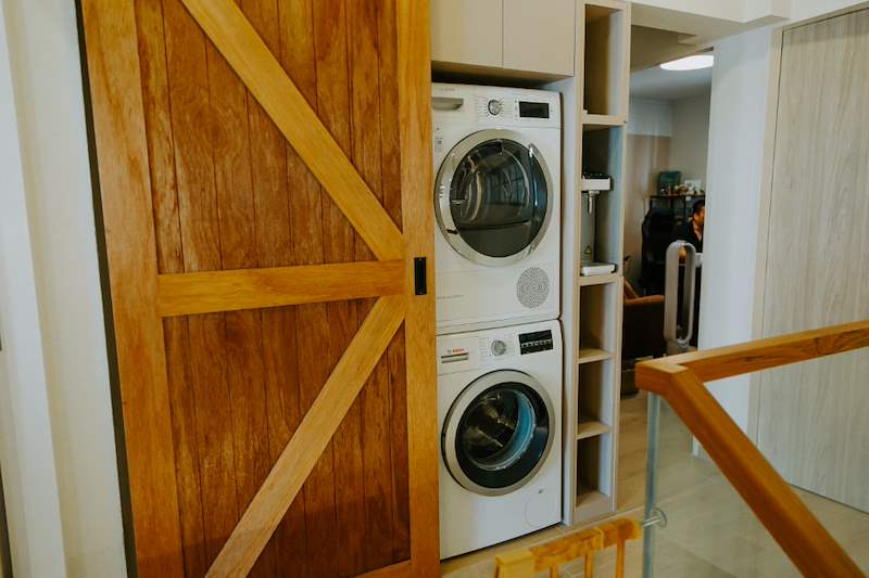 Timber sliding barn door open to a stacked washer and dryer in a laundry cupboard - Full HDB maisonette renovation with a curved timber-slat island and lit staircase (140BB) | Larry Contractors, HDB Licensed Renovation Contractor, Singapore