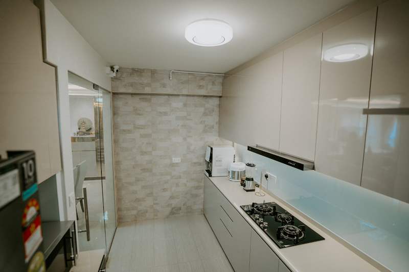 Galley kitchen with cream high-gloss cabinets, a glass splashback, gas hob and a stone-look feature wall at the end - HDB flat renovation in cream and mirror-trim laminate with a galley kitchen (C10) | Larry Contractors, HDB Licensed Renovation Contractor, Singapore