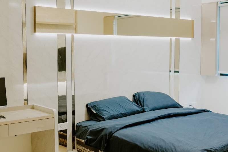 Bedroom with mirror-strip wall panels, a backlit floating shelf and a blue-covered bed - HDB flat renovation in cream and mirror-trim laminate with a galley kitchen (C10) | Larry Contractors, HDB Licensed Renovation Contractor, Singapore