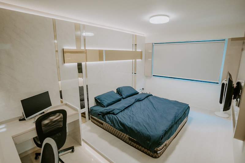 Bedroom with a blue-covered bed, built-in desk, roller blind and cream laminate wardrobes - HDB flat renovation in cream and mirror-trim laminate with a galley kitchen (C10) | Larry Contractors, HDB Licensed Renovation Contractor, Singapore