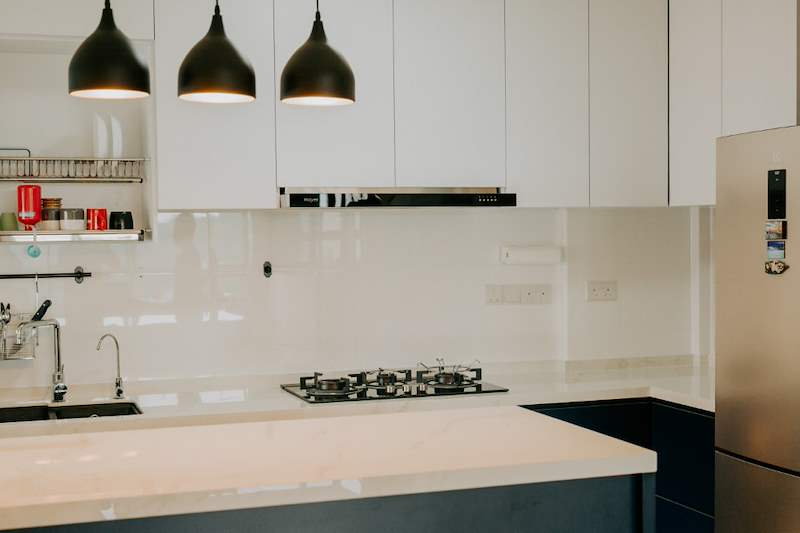 Kitchen with white upper cabinets, glass splashback, gas hob and black pendant lights over the counter - HDB flat renovation with a glass-partitioned study, hidden shoe cabinet and navy kitchen (442BB) | Larry Contractors, HDB Licensed Renovation Contractor, Singapore
