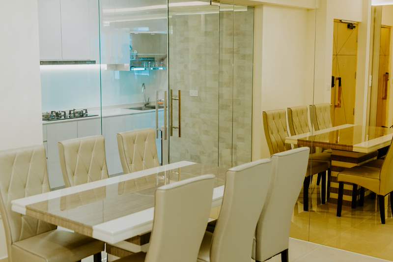 Dining table with cream upholstered chairs beside the glass-walled kitchen - HDB flat renovation in cream and mirror-trim laminate with a galley kitchen (C10) | Larry Contractors, HDB Licensed Renovation Contractor, Singapore