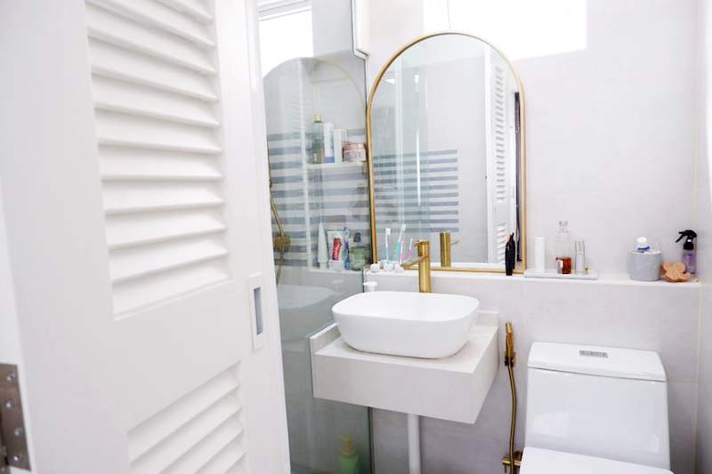 Bathroom with a white countertop basin, brass tap, an arched brass-framed mirror and a louvred door - HDB flat renovation in white shaker-style carpentry with brass handles (C05) | Larry Contractors, HDB Licensed Renovation Contractor, Singapore