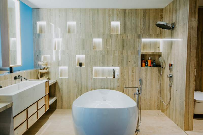 After: bathroom with a freestanding tub, travertine-look walls, lit shelf niches and a rain shower - Condo apartment renovation with a limestone feature wall and dark green kitchen (38MR) | Larry Contractors, HDB Licensed Renovation Contractor, Singapore