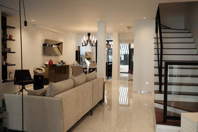 After: open-plan living area with a beige sofa, polished marble-look flooring and a black-framed staircase beyond - Terrace house renovation with new facade and full interior (3TBA) | Larry Contractors, HDB Licensed Renovation Contractor, Singapore