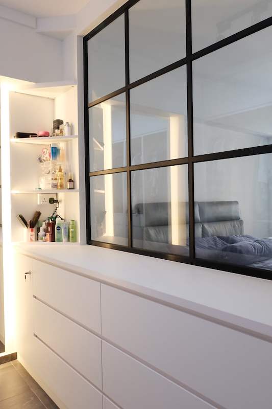 Black-framed glass partition above white drawers, lit shelves in the wardrobe beyond - HDB flat renovation with a navy kitchen and a glass-walled walk-in wardrobe (407B) | Larry Contractors, HDB Licensed Renovation Contractor, Singapore