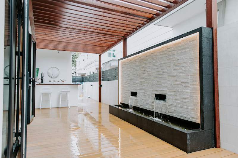 Covered timber deck with a slatted timber ceiling and a backlit stone water-feature wall, outdoor bar behind - Landed house renovation with new facade, staircase and interiors (39EW) | Larry Contractors, HDB Licensed Renovation Contractor, Singapore