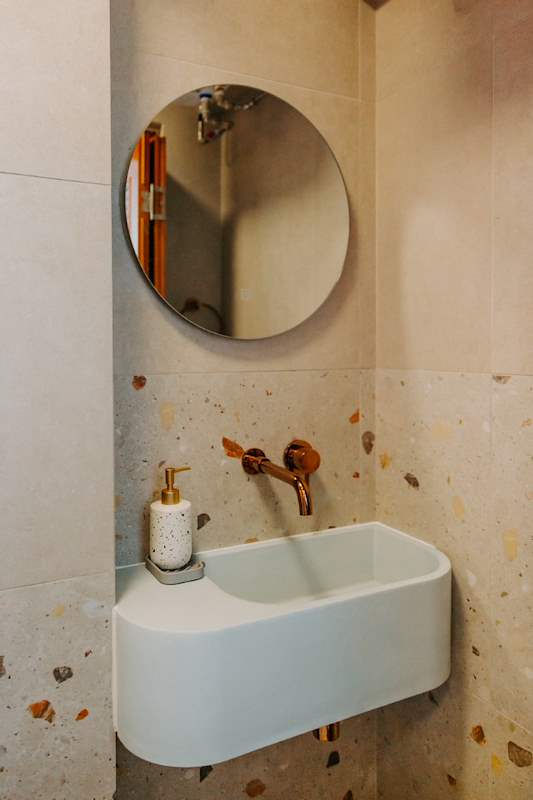 Round mirror above a small wall-hung basin with a brass tap on terrazzo-look tiles - Full HDB maisonette renovation with a curved timber-slat island and lit staircase (140BB) | Larry Contractors, HDB Licensed Renovation Contractor, Singapore