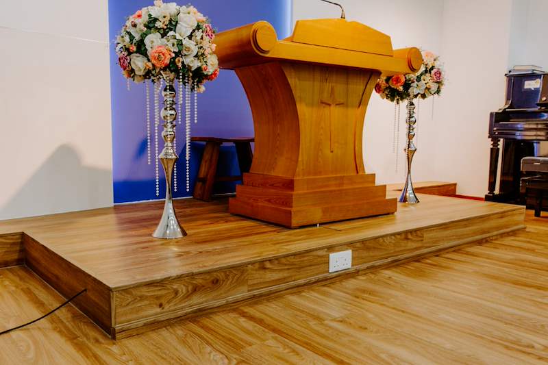 Custom timber podium on the raised timber stage with flower stands and a piano to the side - Church hall fit-out with a timber stage and custom podium (62JBCC) | Larry Contractors, HDB Licensed Renovation Contractor, Singapore