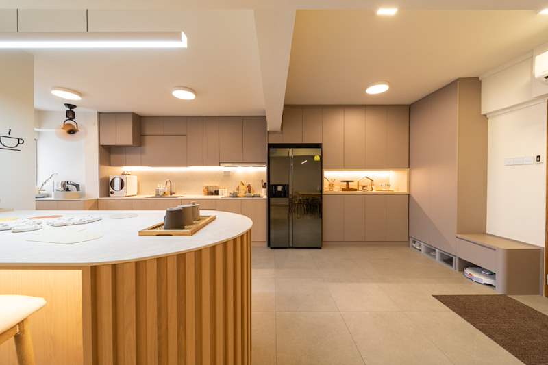 After: the kitchen island and beige cabinets seen from the dining side, fridge and pantry beyond - Full HDB maisonette renovation with a curved timber-slat island and lit staircase (140BB) | Larry Contractors, HDB Licensed Renovation Contractor, Singapore